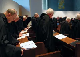 A group of Italian monks in black robes stand in a church, heads bowed over open prayer books.