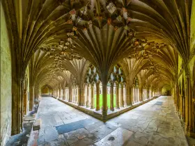 A grand cloister with vaulted ceilings, reminiscent of a monastery floor plan. Sunlight streams through arched windows onto the stone floor.