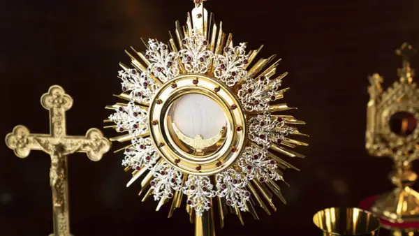 A golden monstrance holds the blessed sacrament, surrounded by ornate details and red jewels. A crucifix stands nearby, a symbol for a blessed sacrament prayer request.
