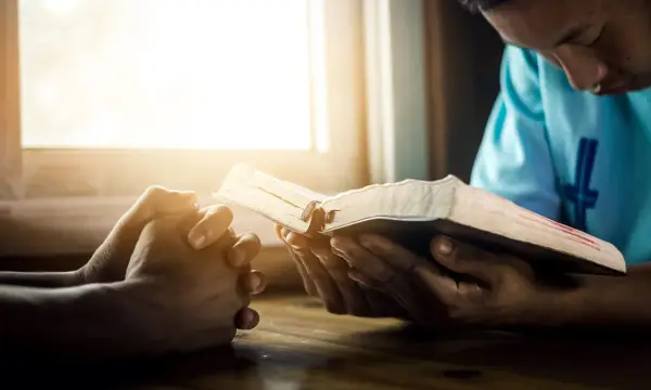 Hands clasped in prayer beside an open Bible, bathed in warm light. A moment of reflection, perhaps contemplating prayer names.