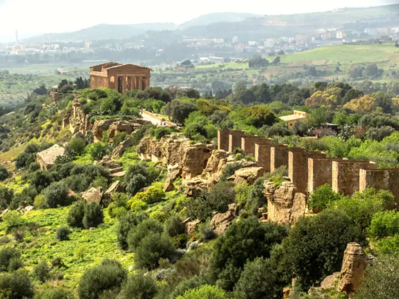 Ancient Greek temples stand proudly on a ridge in the Valley of the Temples, Sicily, surrounded by lush greenery and a distant town.