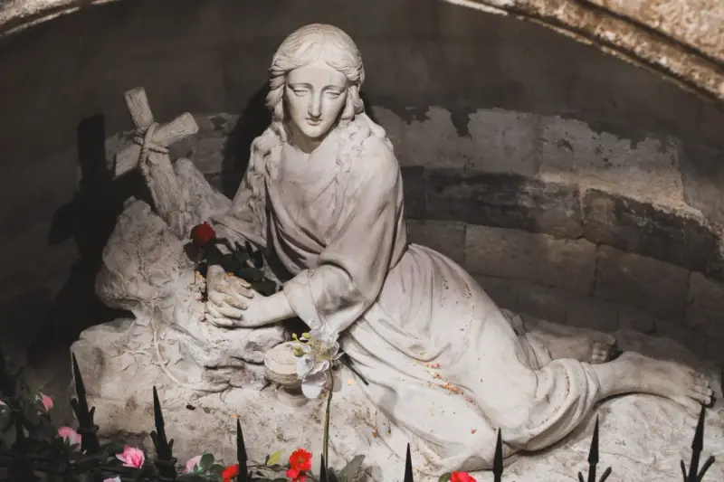 A stone sculpture depicts the serene body of Mary Magdalene, resting with a cross and a single red rose.